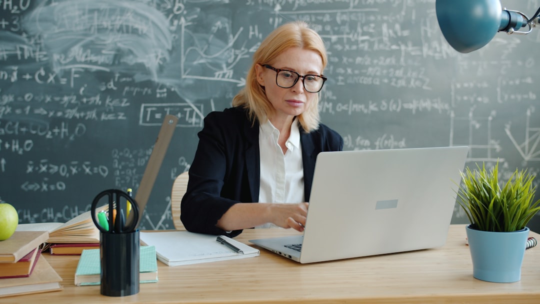 University professor serious adult lady in suit and glasses is using laptop then taking notes working at desk in class. Technology and education concept.