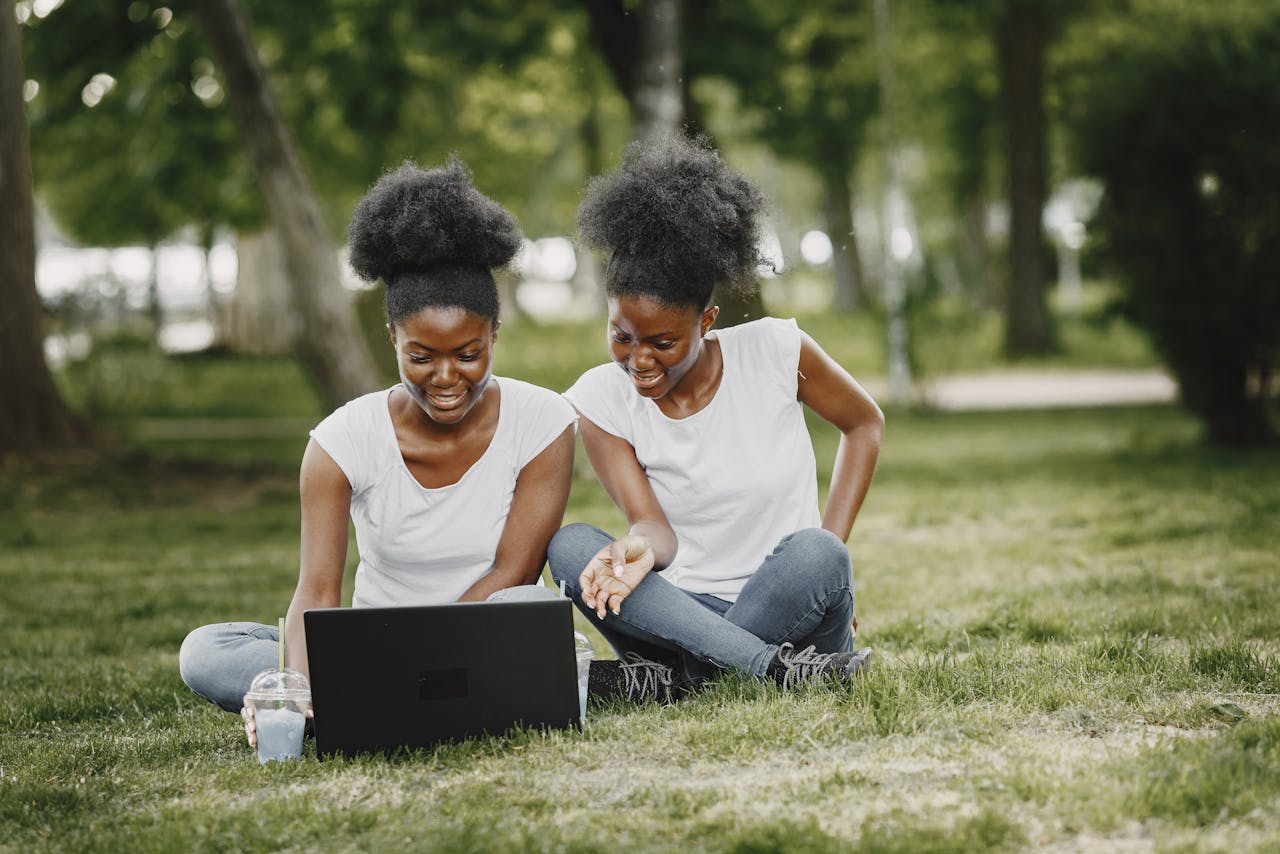 Two women enjoying time with a laptop outdoors, smiling and sitting on grass.