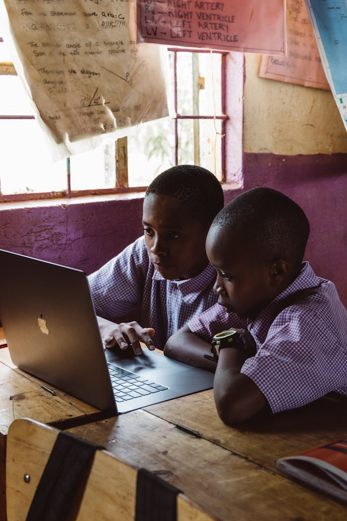 Two young boys in school uniforms focus on a laptop, studying together indoors.